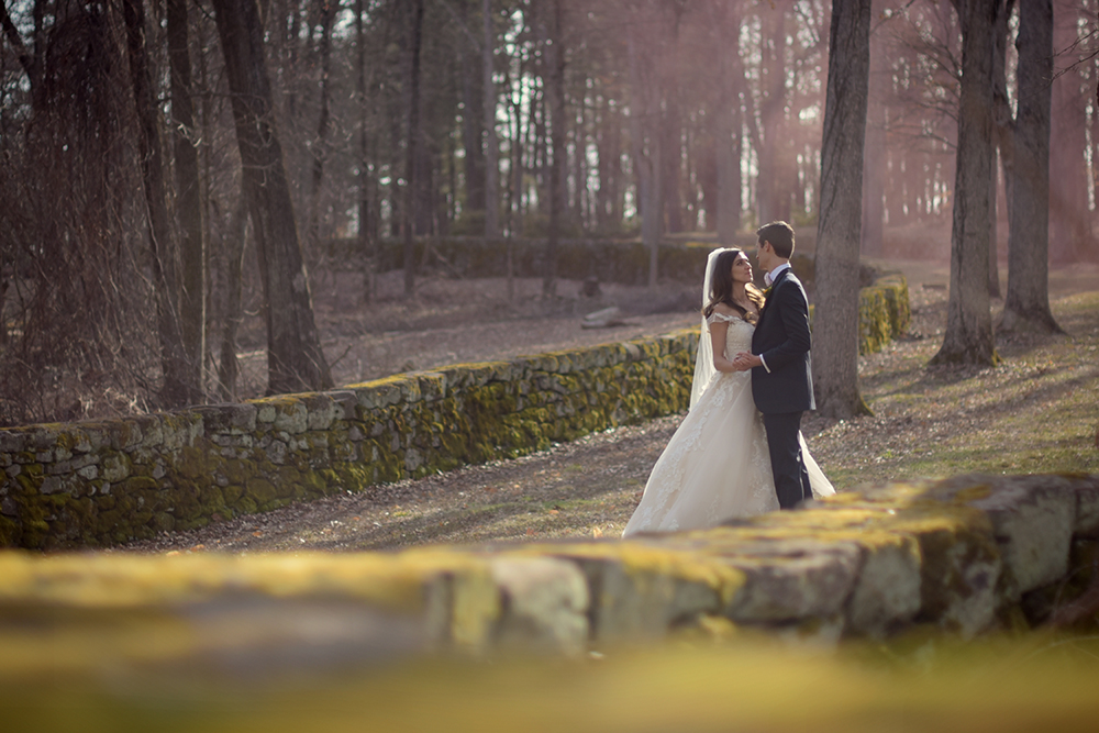 Bride and groom look at each other with trees and stone wall in the background